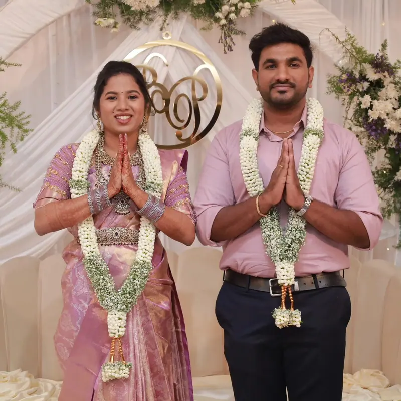 Couple in garlands with folded hands in front of a floral backdrop
